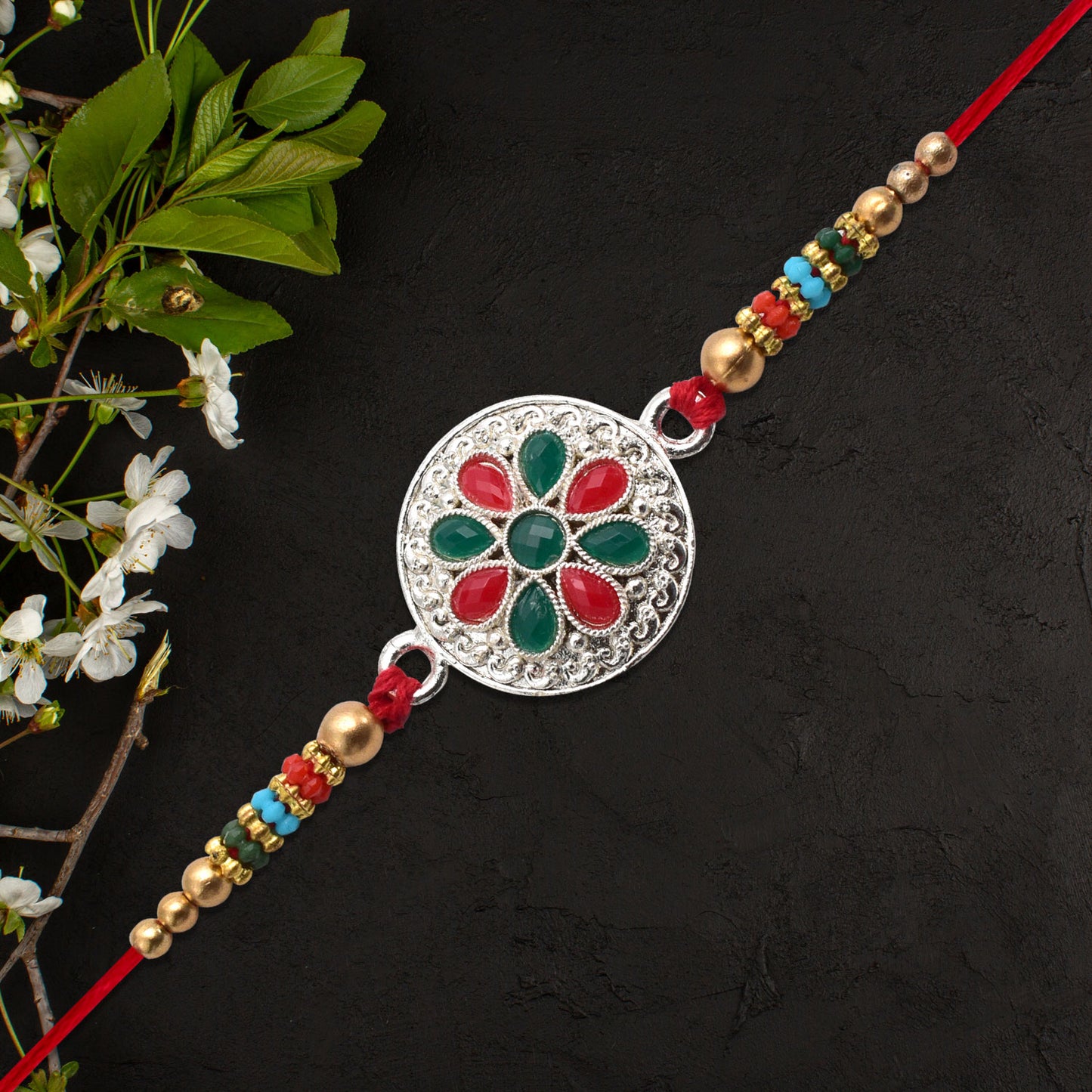 Silver plated floral rakhi with red and green stones, featuring intricate design and vibrant gemstone accents.
