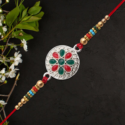 Silver plated floral rakhi with red and green stones, featuring intricate design and vibrant gemstone accents.